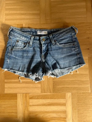 Blå jeansshorts från Lee - Snygga blå jeansshorts från Lee med fransiga kanter. Shortsen har låg midja och slitna detaljer. Perfekta för varma sommardagar. Står inte storlek men skulle säga xs till s.