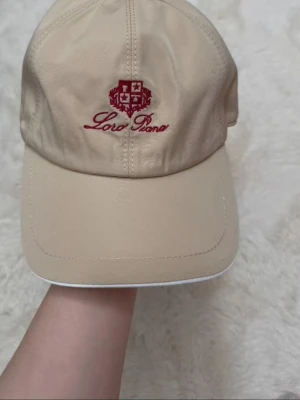 Beige keps från Loro Piana - Stilren beige keps från Loro Piana med röd broderad logotyp och text framtill. Klassisk böjd skärm och justerbar baksida. Tillverkad i ett mjukt, lätt material som ger en exklusiv känsla. Perfekt accessoar för en avslappnad men trendig look.