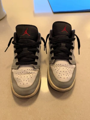 Nike Air Jordan 1 Low grå/vit - Säljer ett par Nike Air Jordan 1 Low sneakers i grått och vitt med svarta detaljer och svart swoosh. Skorna har svart snörning, röd Jumpman-logga på plösen och klassisk låg siluett. Ovandelen är i läder och sulan är vit med svart yttersula.