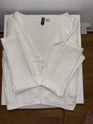 Vit ribbad kofta från Cubus - Säljer en vit ribbad kofta med lång ärm och v-ringad hals. Koftan har knäppning framtill med små vita knappar och är tillverkad i ett mjukt, stretchigt material. Perfekt att slänga över en topp för en enkel och clean look.