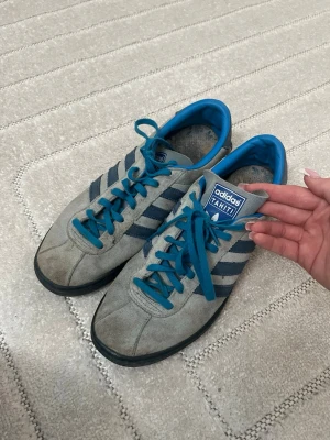 Gamla Adidas Tahiti sneakers - Säljer ett par Adidas Tahiti sneakers i grå mocka med blå detaljer, blå ränder och blå snörning. Skorna har klassisk låg profil och svart sula. Perfekta för dig som gillar retrostil och vill ha något unikt från Adidas Originals. 
