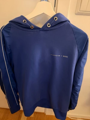 Blå hoodie Porsche x Boss - Snygg blå hoodie från Porsche x Boss med vita detaljer och logga på bröstet. Hoodien har huva med dragsko och metallöljetter samt vita ränder längs ärmarna. Tillverkad i mjukt material som känns skönt mot huden. Perfekt för en sportig och stilren look. Finns litet hål på armen men det syns inte när man har på sig den och det är blått tyg bakom så inget ses igenom