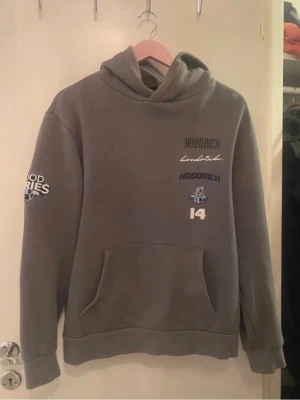 Grå hoodie från Hoodrich med tryck - Snygg grå hoodie från Hoodrich med flera tryck och broderier på bröstet och ärmen. Klassisk känguruficka framtill och huva. Perfekt för en streetwear-look och passar till jeans eller joggers.