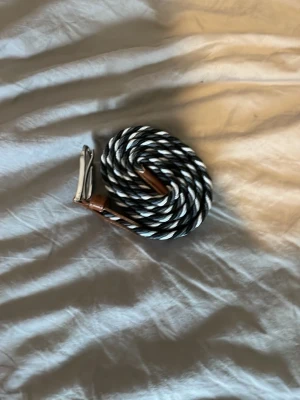 Randigt flätat bälte med läderdetaljer - Snyggt flätat bälte i svart och vitt med randigt mönster. Bältet har silverfärgat spänne och bruna läderdetaljer vid spännet. Perfekt accessoar för att lyfta din outfit med en cool touch.