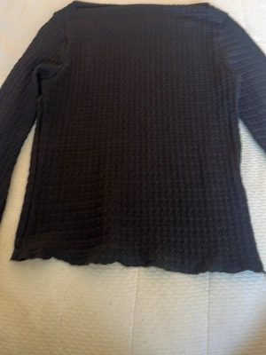 Svart långärmad topp med struktur - Säljer en svart långärmad topp med vågig struktur i tyget. Tröjan har en rak passform och är tillverkad i ett stretchigt material som ger en cool textur. Perfekt för dig som gillar stilrena plagg med det lilla extra.