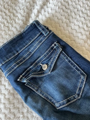 Blå jeans från Gina Tricot - Snygga blå jeans från Gina Tricot med vita kontrastsömmar och klassiska fickor. Jeansen har en knappstängning och bälteshällor, perfekt för en trendig och avslappnad look. Materialet är denim i bomull med lite stretch för extra komfort.