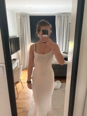 Lång vit klänning med smala axelband - Säljer en fin långklänning i beige med smala axelband och figurnära passform. Klänningen har en enkel och stilren design som framhäver siluetten. Perfekt för dig som vill ha en klassisk look.
