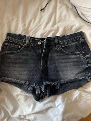 Svarta jeansshorts med fransar - Säljer ett par svarta jeansshorts med råa franskantade benslut och klassisk femficksdesign. Shortsen har en låg midja och slitningar på toppen.