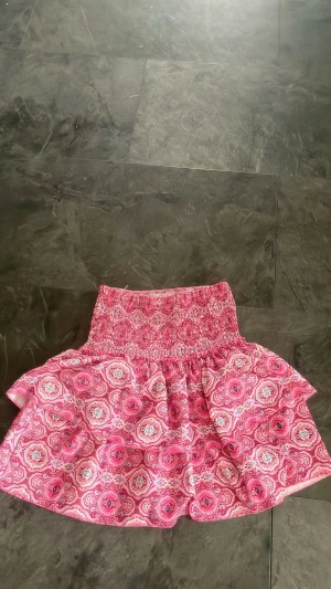 Rosa mönstrad kortkjol med volang - Supersöt rosa kortkjol med bred smock i midjan och volangig kjoldel. Kjolen har ett paisleyinspirerat mönster i vitt och rosa samt ett vitt innerfoder för extra komfort. Perfekt för dig som vill ha en lekfull och färgglad stil.