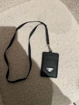 Svart korthållare  - Snygg svart korthållare i läder med avtagbart band för att hänga runt halsen. Framsidan har en ikoniska metallogo och baksidan har ett genomskinligt fack för ID eller busskort. Perfekt för dig som vill ha koll på korten med stil.