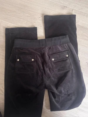 Juicy Couture mjukisbyxor i velour - Säljer ett par svarta mjukisbyxor i mjuk velour med resårmidja och dragsko. Byxorna har två fickor fram och två bakfickor med lock och silverfärgade knappar. Diskret broderad logga på ena fickan framtill. Perfekta för chill dagar.