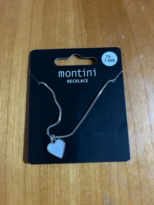 Halsband med hjärthänge från Montini - Snyggt halsband i silverfärg från Montini med ett stilrent hjärthänge. Kedjan är tunn och elegant, perfekt för att ge en söt touch till din outfit. Hjärtat har en slät yta och halsbandet är enkelt att matcha med andra smycken.