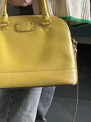 Gul handväska från Kate Spade - Snygg gul handväska från Kate Spade i slätt läder. Väskan har två handtag, guldfärgade detaljer och dragkedja upptill. Insidan är fodrad med mönstrat tyg och har innerfack för småsaker. Perfekt till våren och sommaren för dig som vill sticka ut med färg. Väskan är i nyskick.