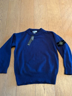Blå stickad tröja från Stone Island - Snygg blå stickad tröja från Stone Island med klassisk rund hals och ribbade muddar. Tröjan har den ikoniska Stone Island-loggan på vänster ärm och är tillverkad i mjukt material. Perfekt för dig som vill ha en stilren och trendig look.