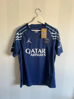 Blå PSG matchtröja Jordan x Nike - Snygg blå PSG matchtröja med vita detaljer och Qatar Airways-tryck på bröstet. Tröjan har korta ärmar, rund hals och PSG-logga samt Jordan-logga framtill. Tillverkad i lätt och ventilerande material, perfekt för fotboll eller träning.