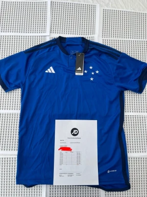 Blå Adidas fotbollströja Cruzeiro - Snygg blå fotbollströja från Adidas med Cruzeiro-logga och fem vita stjärnor på bröstet. Tröjan har korta ärmar, rund hals och mörkblå detaljer på axlar och sidor. Tillverkad i lätt och ventilerande material, perfekt för träning eller match.