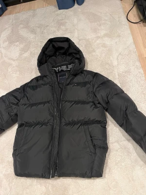 Svart pufferjacka från Tommy Hilfiger - Nypris: 2 000kr. Snygg svart pufferjacka från Tommy Hilfiger med huva (avtagbar) och dragkedja. Jackan har broderad logga på insidan av huvan och diskret flagga på bröstet. Tillverkad i slitstarkt material och fodrad för extra värme. Perfekt för kalla vinterdagar. Köp på Kids Brandstore för nått år sedan och använde den vara några fåtal gånger. 