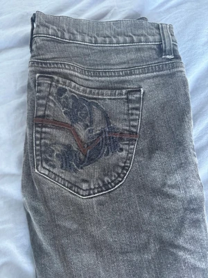 Grå bootcut jeans med broderad ficka - Säljer ett par grå bootcut jeans med snygg broderad bakficka och en brun detaljsöm. Jeansen har slitsar nedtill på benen och klassisk femficksdesign. Perfekta för dig som gillar en avslappnad men ändå trendig stil.