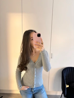 Ljusgrå ribbad kofta med v-ringning - Ljusgrå ribbad kofta med v-ringning och knappar framtill. Figurnära passform och långa ärmar. Perfekt att styla med jeans för en enkel och snygg look. Materialet ser ut att vara mjukt och stretchigt stickat tyg.