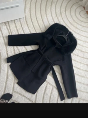 Svart kappa med päls och skärp - Stilren svart kappa med stor krage i päls och knytband i midjan. Kappan har stora fickor framtill och en rak passform som ger en snygg siluett. Perfekt för kyliga dagar när du vill vara både varm och trendig.