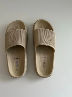 Beige sandaler / tofflor från CUSHIONAIRE i storlek 44 - Säljer ett par stilrena beige tofflor från CUSHIONAIRE i storlek 44. De har en bred ribbad rem över foten och är gjorda i ett mjukt, flexibelt material som känns skönt att gå i. Perfekta för att chilla hemma eller på stranden.