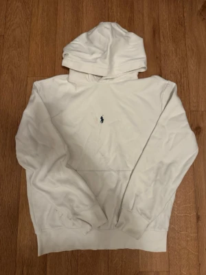 Vit hoodie från Polo Ralph Lauren - Klassisk vit hoodie från Polo Ralph Lauren med broderad marinblå logga på bröstet. Hoodien har huva, känguruficka och ribbade muddar vid ärmslut och nederkant. Tillverkad i mjukt bomullsmaterial för en skön och avslappnad look.