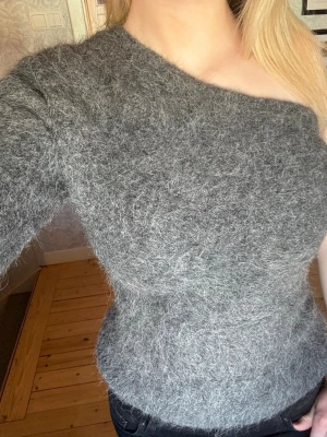 Grå off shoulder stickad tröja - Säljer en fluffig grå stickad tröja med off shoulder-design och en ärm. Tröjan har en kroppsnära passform och är gjord i ett mjukt, lurvigt material som ger en mysig känsla. Perfekt för dig som vill ha något unikt och trendigt i garderoben.