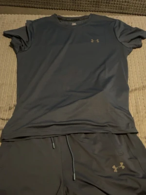 Mörkblått träningsset från Under Armour - Snyggt mörkblått träningsset från Under Armour med t-shirt och shorts. Tillverkat i ett lätt och snabbtorkande funktionsmaterial som andas. T-shirten har rund hals och korta ärmar, medan shortsen har dragsko i midjan och logga på benet.