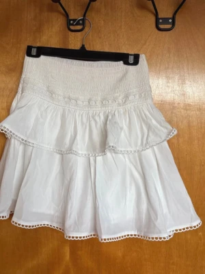 Vit volangkjol med spetsdetaljer - Supersöt vit kjol med volanger och fina spetsdetaljer längs kanterna. Kjolen har en smockad midja som ger en bekväm passform och ett luftigt fall. Perfekt för varma dagar och ger en romantisk vibe till din outfit. Modellen är Onesize och sitter jättefint oavsett om man vill bära den högt i midjan eller lite längre ner på höften. 