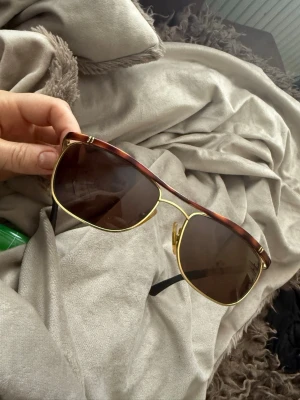 Bruna aviator-solglasögon från Police - Snygga aviator-solglasögon från Police med bruna glas och guldfärgade detaljer. Bågarna har en brun marmorerad look och metallinslag som ger ett exklusivt intryck. Perfekt för dig som vill ha en cool och klassisk stil.