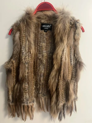 Brun pälsväst från Hollies - Säljer en fluffig och lyxig pälsväst från Hollies i olika bruna nyanser. Västen har en öppen front och är gjord av mjuk päls med längre detaljer nedtill. Perfekt för dig som vill ha en statement piece till din outfit.