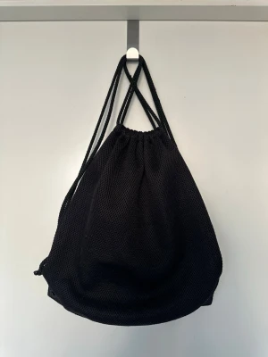 Svart gympåse i mesh - Säljer en svart gympåse i meshmaterial med dragskostängning och tunna axelband. Väskan har en enkel och sportig design, perfekt för träning eller att bära med sig det viktigaste. Lätt och smidig att ta med överallt.