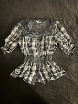 Blårutig blus med trekvartsärm - Snygg blårutig blus med trekvartsärmar och knappar framtill. Blusen har en markerad midja med resår och volang nedtill. Kragen är bred och rundad med en kontrast i enfärgat tyg. Perfekt för en avslappnad men trendig look.