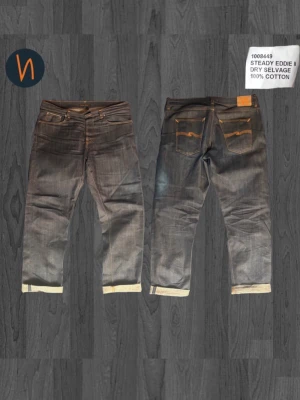 Nudie Jeans Steady Eddie II Selvedge - Säljer ett par mörkblå Steady Eddie II Dry Selvedge jeans från Nudie Jeans Co. Klassisk rak passform med orange kontrastsömmar och vikta benslut. Tillverkade i 100% bomull med rå, otvättad denim för en autentisk look.