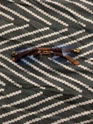 Blåtonade solglasögon med trädetaljer - Unika solglasögon med blåtonade glas, guldfärgade metalldetaljer och snygga skalmar i trä. Glasögonen har en modern, kantig form och ger en cool vibe till din stil. Perfekt accessoar för dig som vill sticka ut.