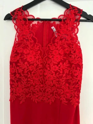 Röd långklänning med spetsdetaljer - Elegant röd långklänning med blommiga spetsdetaljer upptill och slits framtill. Klänningen har ärmlös design och rund halsringning. Perfekt för speciella tillfällen där du vill sticka ut.