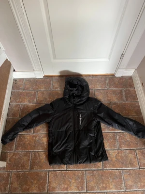 Svart sailracing pufferjacka med huva - Snygg svart pufferjacka med huva och dragkedja framtill. Jackan har en ficka med dragkedja på bröstet och två sidofickor. Tillverkad i glansigt material som ger en modern look. Perfekt för kalla dagar när du vill hålla dig varm och stilren.