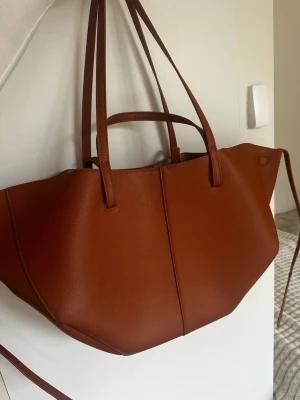 Brun shoppingväska i konstläder - Stilren shoppingväska i brunt konstläder med geometrisk form och dubbla handtag. Väskan har en modern design med släta ytor och långa axelband, perfekt för dig som vill ha plats för allt. Enkel att matcha till olika outfits.