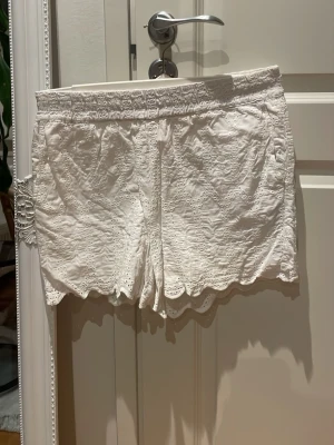 Vita broderade shorts med spetskant i storlek 36 - Säljer ett par vita shorts från Amisu med broderade detaljer och spetskant. De har elastisk midja och är tillverkade i ett lätt bomullstyg, perfekta för varma dagar. Shortsen har en feminin look med fina hålbroderier och vågig nederkant.