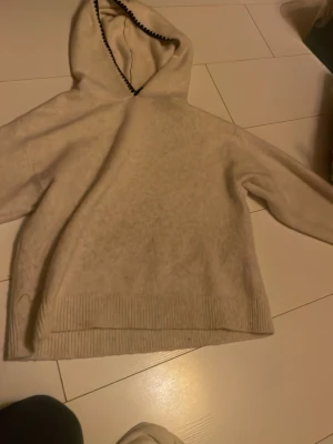 Beige hoodie med ribbade detaljer - Mysig beige hoodie med huva och ribbade muddar vid nederkant och ärmslut. Huvan har en dekorativ mörk söm längs kanten. Perfekt för en avslappnad och stilren look. Materialet ser ut att vara mjukt och lite luddigt, vilket ger en skön känsla.