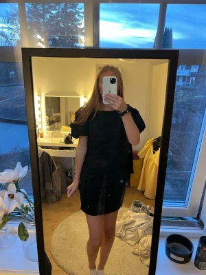 Svart kort klänning med puffärm - Snygg svart kort klänning med puffärmar och öppen rygg. Klänningen har en enkel och puffig passform och är tillverkad i ett slätt tyg som ger en stilren look. Perfekt för dig som gillar minimalistisk stil med en twist. Dragkedja finns på sidan!