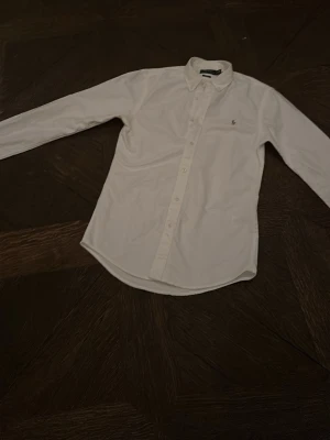 Vit skjorta från Ralph Lauren - Klassisk vit skjorta från Ralph Lauren med button-down krage och broderad logga på bröstet. Skjortan har lång ärm och knäppning framtill. Perfekt för en clean och stilren look.