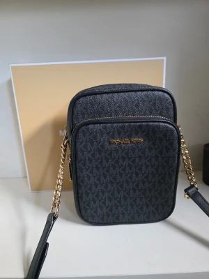 Svart axelväska Michael Kors - Snygg svart axelväska från Michael Kors med guldiga detaljer och kedjerem. Väskan har MK-logga över hela materialet och två fack med dragkedja. Tillverkad i tåligt konstläder och perfekt för dig som vill ha en stilren och trendig accessoar.