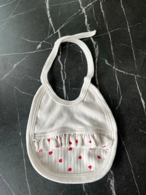 Vit haklapp med röda hjärtan - Söt vit haklapp med knytband och en ficka framtill dekorerad med små röda hjärtan. Tillverkad i mjukt tyg som känns bekvämt mot huden. Perfekt accessoar för att hålla kläderna rena under måltider.