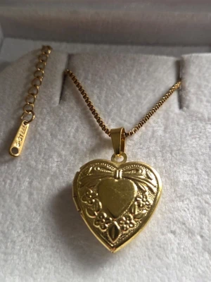 Guldigt hjärthalsband med medaljong - Säljer ett guldtonat halsband i rostfritt stål med en hjärtformad medaljong. Medaljongen har fina blommönster och en liten rosett på framsidan, och går att öppna för att lägga in ett foto eller minne. Kedjan är tunn och elegant, perfekt för att ge en personlig touch.