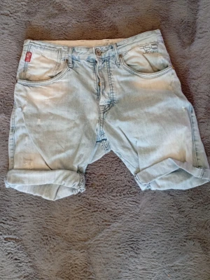 Ljusblå jeansshorts  - Säljer ett par ljusblå jeansshorts med klassisk femficksdesign och uppvikta benslut. Shortsen har knappgylf och detaljerade sömmar på bakfickorna. Perfekta för varma dagar och enkel att matcha med olika stilar.