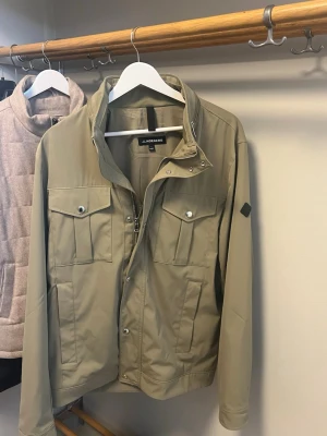Beige vindjacka från J.Lindeberg - Stilren beige vindjacka från J.Lindeberg med dragkedja och tryckknappar framtill. Jackan har två bröstfickor med lock och två sidofickor. Långärmad modell i ett lätt och smidigt material, perfekt för vår och höst.