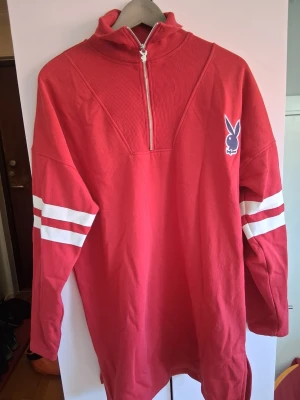 Röd långärmad tröja från Playboy - Röd långärmad tröja från Playboy med vit dragkedja vid halsen, vita ränder på ärmarna och stort Playboy-tryck på ryggen. Tröjan har hög krage och är tillverkad i mjukt bomullsmaterial. Perfekt för en avslappnad streetwear-look.