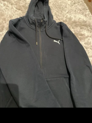 Mörkblå hoodie från Puma - Snygg mörkblå hoodie från Puma med dragkedja framtill och vit Puma-logga på bröstet. Hoodien har huva med dragsko och en stor magficka. Perfekt för en avslappnad och sportig stil.