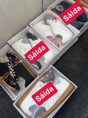 Svarta sneakers med färgstänk - Unika svarta sneakers från Maison Margiela med färgstänk i rött, blått, gult och vitt. Skorna har vita snören, brun sula och en klassisk låg siluett. Perfekta för dig som vill sticka ut med din stil.